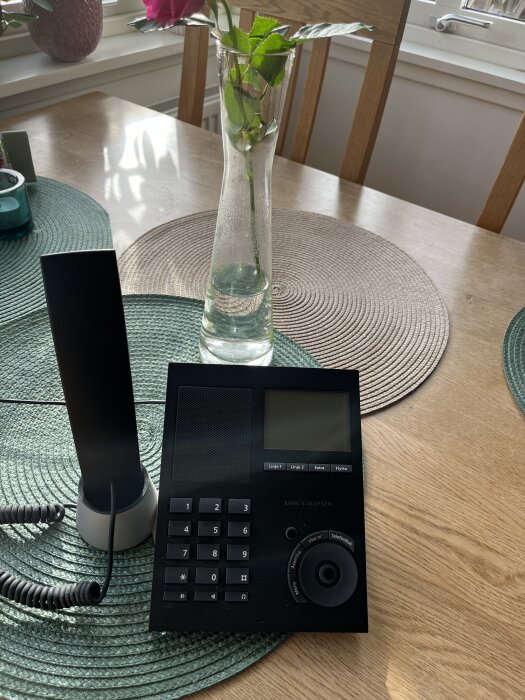 Trådlös telefon och vas med ros på ett träbord med runda bordstabletter i soligt rum.