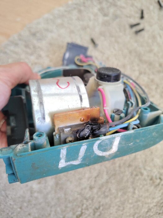 Elektrisk komponent med sladdar och märken, skadad med brännmärken, hålls i en hand över en matta.