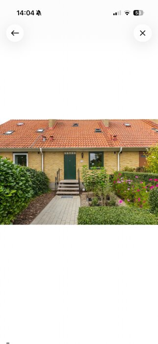 Radhuslänga med gul tegel, rött taktegel och grön dörr, omgiven av grönska och blommor.