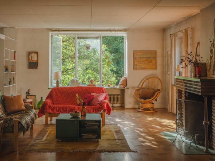 Ljust vardagsrum i fabrikörsvilla från 1946 med stora fönster, röd soffa, fåtölj, öppen spis och bokhylla.