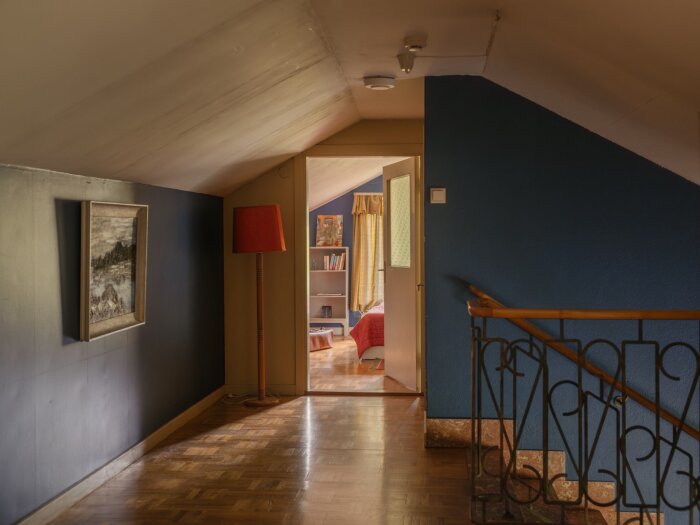 Övre hall i en fabrikörsvilla med snedtak, blå väggar, röd golvlampa och tavla, leder in till ett sovrum med guldfärgade gardiner.