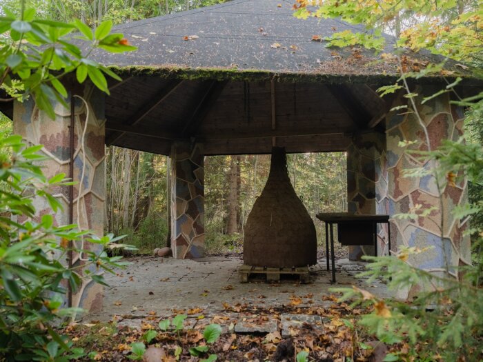 Paviljong med sexkantigt tak och färgglatt mosaikmönster på pelarna, omgiven av träd. Inuti syns ett bord och en dekorativ struktur.