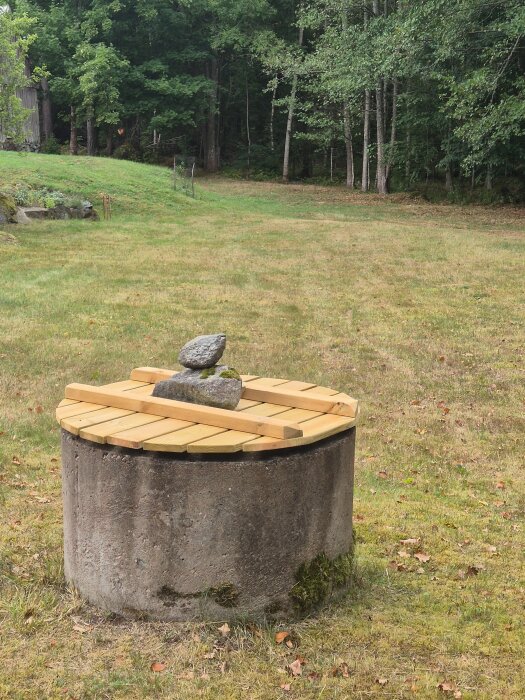 Betongbrunn med trälock och stenar ovanpå, omgiven av gräs och skog i bakgrunden.