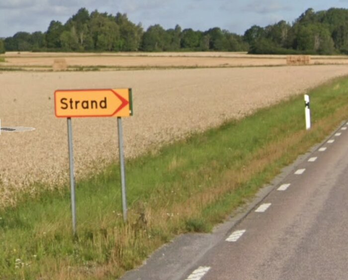Vägskylt som visar riktning mot "Strand" vid en landsväg och åker.