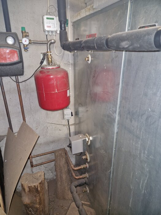 Gammalt pannrum med röd tryckexpansionskärl, rör, manometer och isolerade rör på en betongvägg. Trästockar och träskivor på golvet.