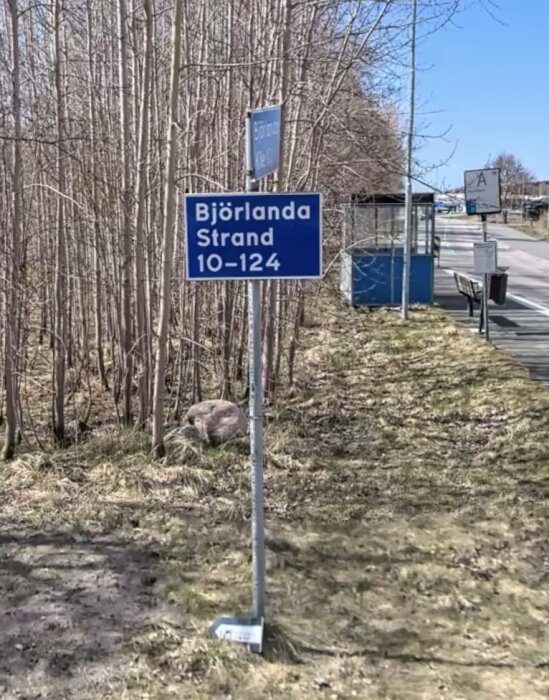 Skylt med texten "Björlanda Strand 10-124" vid en vägkant omgiven av träd och en busshållplats i bakgrunden.