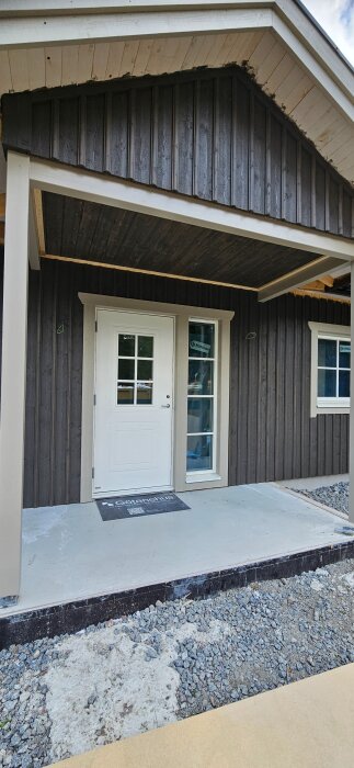 Förstukvist med betongplatta framför trädörr, under ett tak med träpanel, omgiven av grus under byggtid.