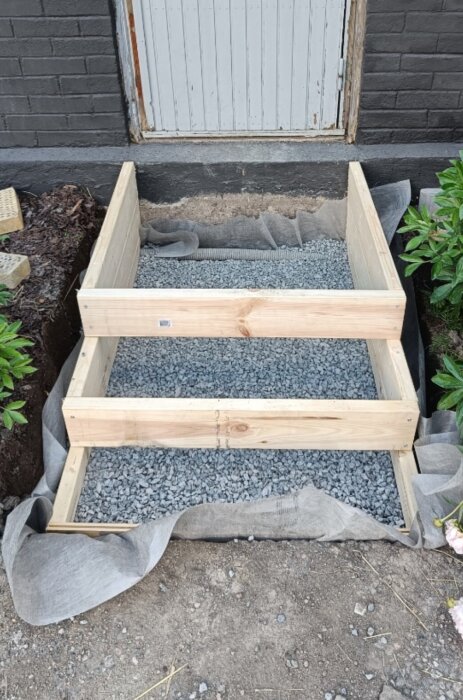 Träram fylld med grus, placerad framför mur och dörr, förberedd för betonggjutning av trappa.
