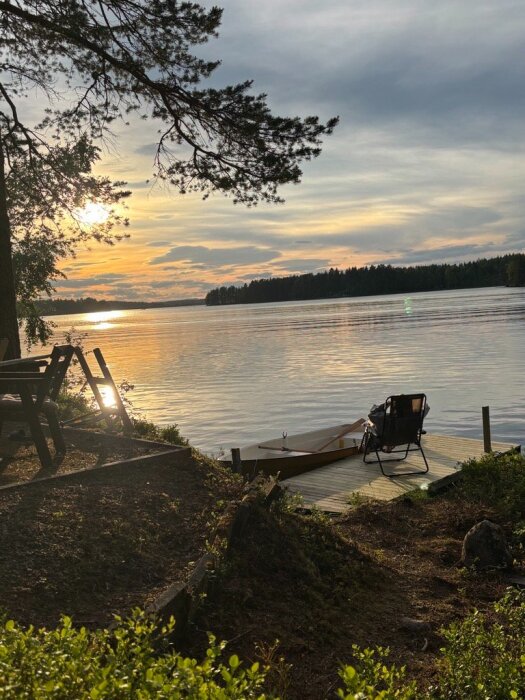 Solnedgång vid sjö med brygga och båt, två stolar står på bryggan bredvid vattnet. Träd och buskar omger stranden.