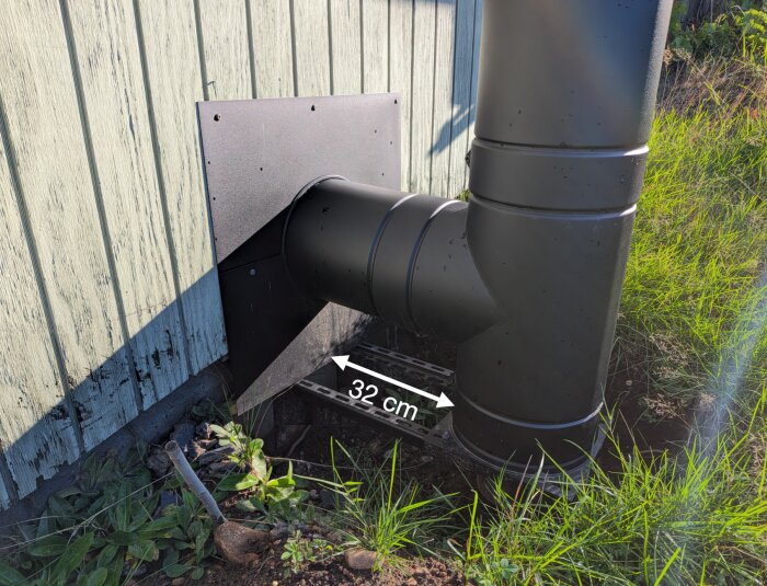 Svart skorsten installerad nära husgrund med vit pil som visar avstånd 32 cm till väggen; omgiven av gräs och jord.