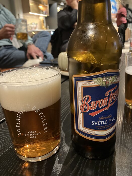 Ölflaska med etiketten "Baron Trenck" och ett glas med öl från Gotlands Bryggeri på ett svart bord, människor i bakgrunden.