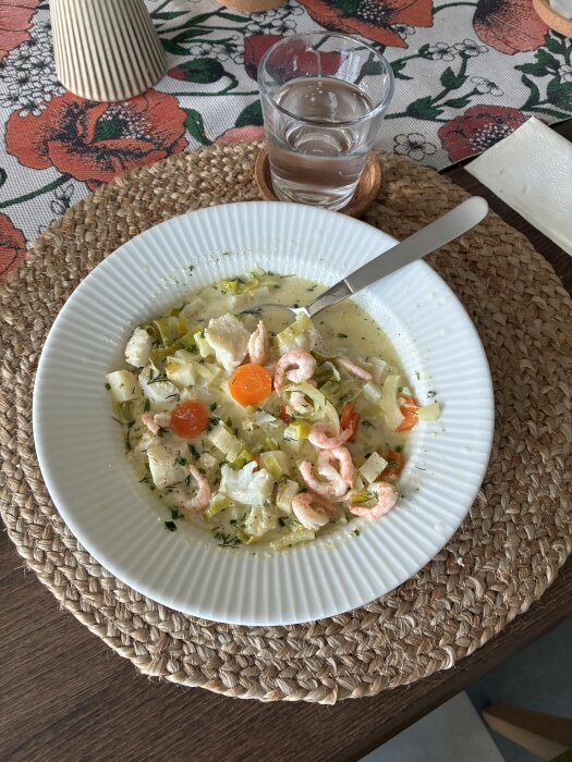 Tallrik med fisksoppa innehållande räkor, morötter, fänkål, purjolök och rotselleri, serverad på bord med glas vatten bredvid.