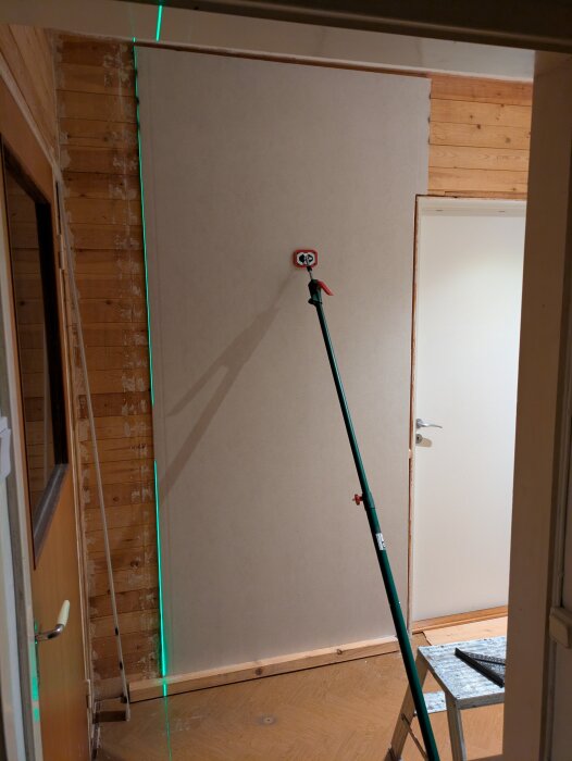 Gipsskiva monterad i hall under renovering, stöd av grön måttstång, synlig träram och laserlinje.