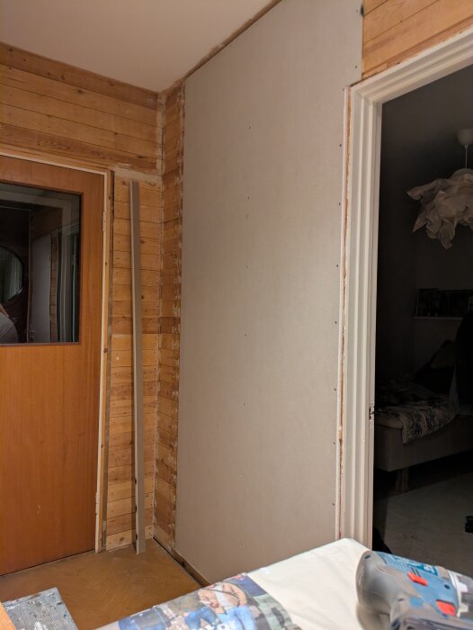 Första gipsskivan monterad i hall, omgiven av träpanel och dörröppningar. Renoveringsarbete med verktyg i förgrunden.