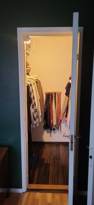 Walk-in closet med dörr öppen, kläder hänger på vänster sida och slipsar på väggen längst bak, mörkt trägolv och vita väggar.