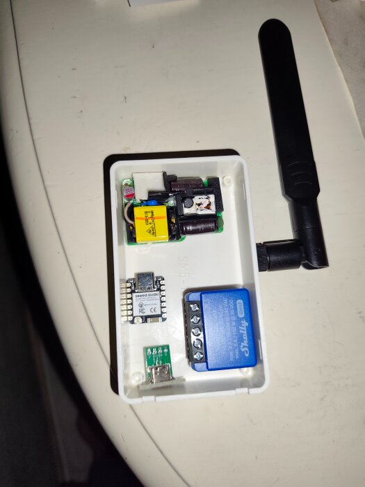 Öppen liten låda med elektroniska komponenter, inklusive en Shelly mini puck, ESP32 C6, panel-USB-uttag, och en antenn, på ett bord.
