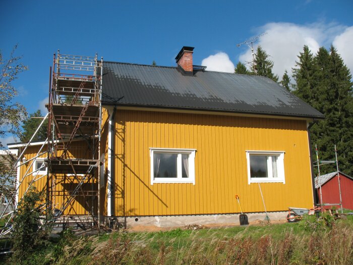Gult hus med svarta takpannor under renovering, byggnadsställningar på sidan. Klar blå himmel, grönska i bakgrunden.