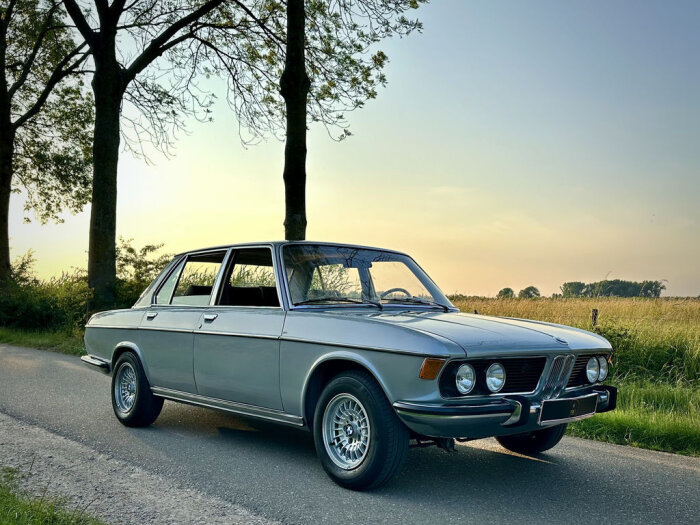 Klassisk BMW E3, även känd som "Storbumsen", parkerad vid vägkant omgiven av träd och gräs i solnedgång.