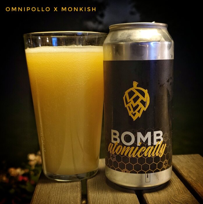 Ölglas med guldröd dryck bredvid en aluminiumburk med texten "Bomb Atomically" och fruktig humlesymbol på ett träbord.