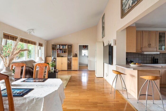 Öppen planlösning med glansig gul-orange parkett, matbord, stolar och kök med ljusa skåp och köksö, samt vitrinskåp i vardagsrum.