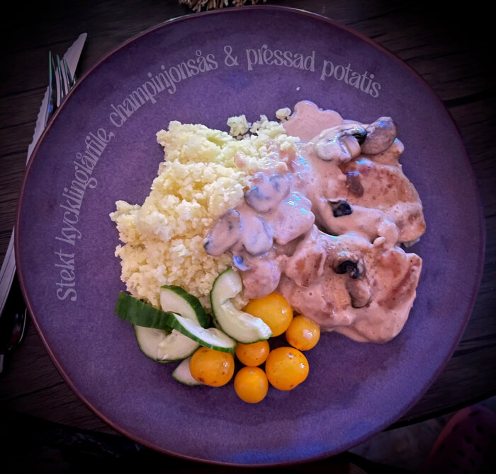 Stekt kycklinglårfilé med champinjonsås, pressad potatis, gurkskivor och gula tomater på en lila tallrik.