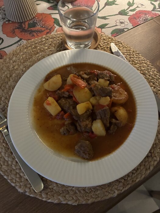 Gryta/gulasch på tallrik med högrev, paprika, lök, chili, vitlök, spiskummin och potatis. Glas med vatten i bakgrunden.