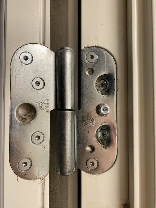 Gångjärn på en dörrkarm med flera synliga skruvar och hål, används för att justera dörrhöjd. Märkt med "ASSA" och installerat år 2011.