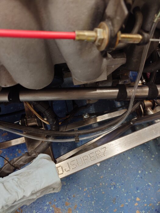 Motorstruktur med rör och kablar, ett röd kabel kopplad till del. Texten "SUPER 7" synlig på metallram i blåfläckig bakgrund.