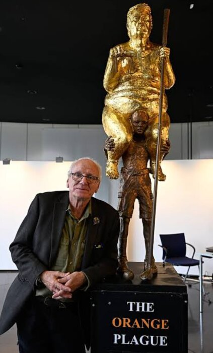 En man står bredvid en skulptur av en större, gyllene figur ovanpå en annan, mindre figur med skylten "The Orange Plague".