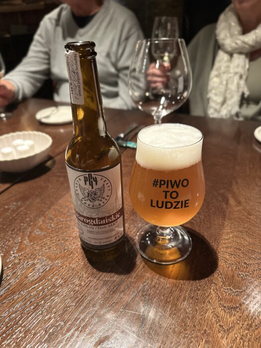 Ölglas med texten "#PIWO TO LUDZIE" bredvid en ölflaska på ett träbord i Gdansk, omgiven av personer med vinglas.