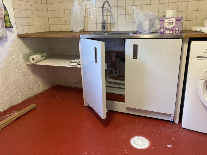 Öppet underskåp med synliga rör under diskbänk i kök, röd golvfärg, pappersrulle på hylla, tvättmedelspaket och soppåse på bänkskiva.