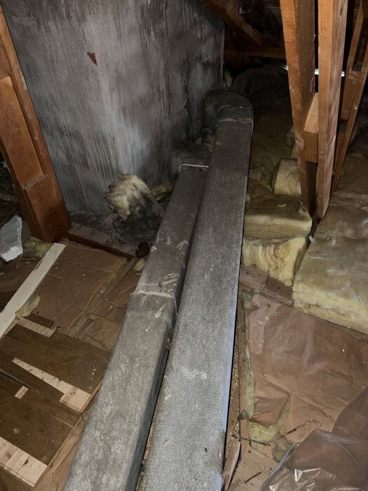 Asbestbelagt ventilationsrör på vinden kopplat till murstock, delvis igentäppt med fågelbo och omgiven av isoleringsmaterial.