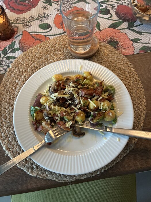 Tallrik med gnocchi, svamp och bacon toppad med riven ost, bredvid ett glas vatten på bord dukat med blommig löpare och ljuslykta.