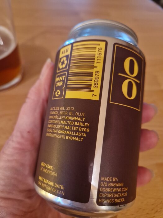 En hand håller en brun ölburk från 0/0 Brewing med gul text och symboler på etiketten.