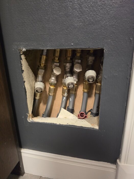 Lucka i vägg med flera kopplingsrör och ventiler, vissa rör märkta med justeringsrattar, en del av hemuppvärmningssystemet.
