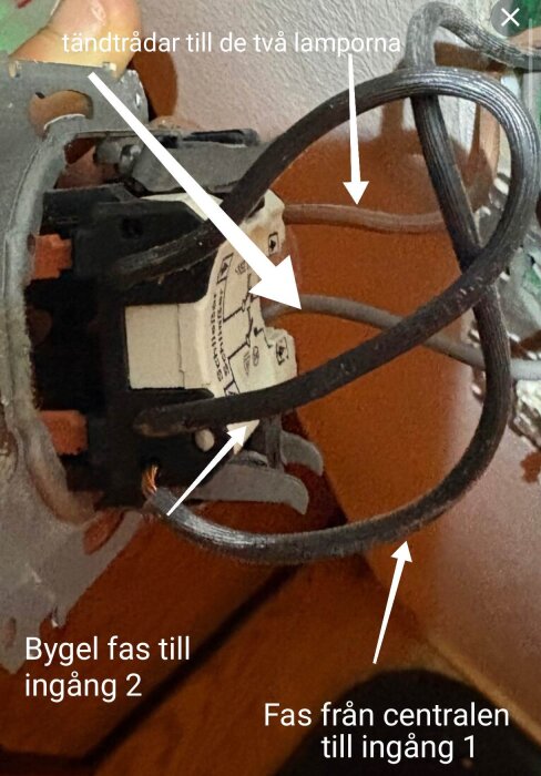 Närbild på elinstallation med ledningar markerade som "tändtrådar till de två lamporna", "fas från centralen till ingång 1" och "bygel fas till ingång 2".