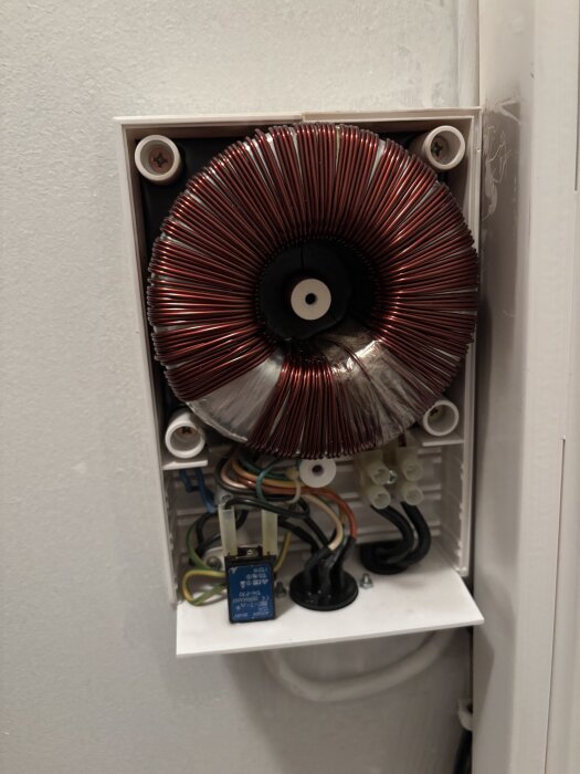 Elektrisk komponent med lindad koppartråd och kablar inuti en vit låda på väggen.