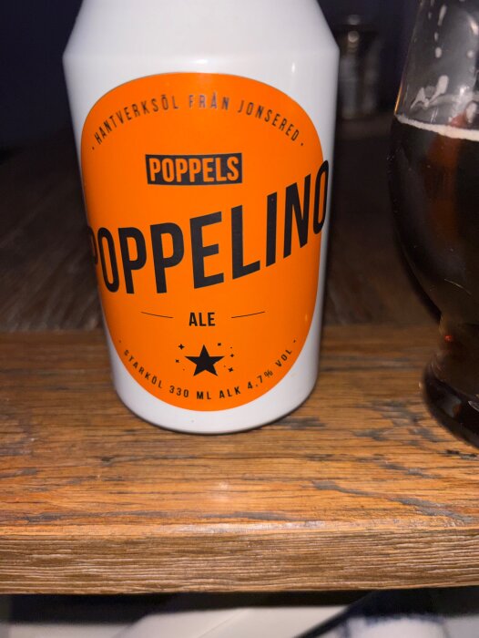Burk med Poppelino ale stående på ett träbord bredvid ett glas öl. Orange etikett med information om bryggeri och alkoholinnehåll.