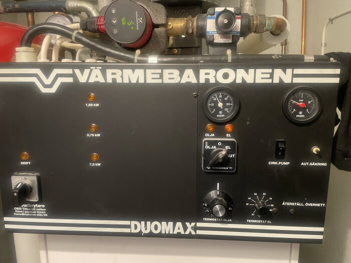 Kontrollpanel med termostat och vridknappar på en Värmebaronen-panna, märken för olja och el, samt en tryckmätare.