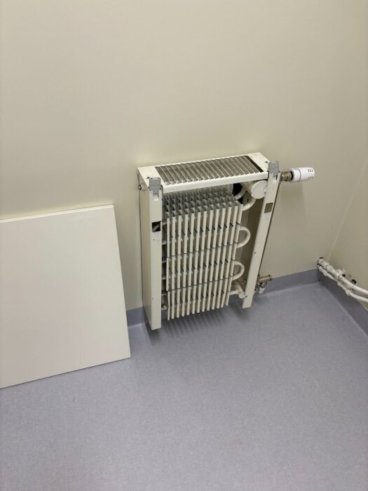 Radiator med borttagen front, visar rörslingor och lameller, installerad i ett rum med ljusa väggar och golv.