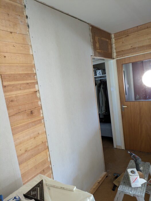 Inre renovering, halvgjort rum med träpaneler och gipsskiva, verktyg och material som borrmaskin och måttstock synliga på en stege.