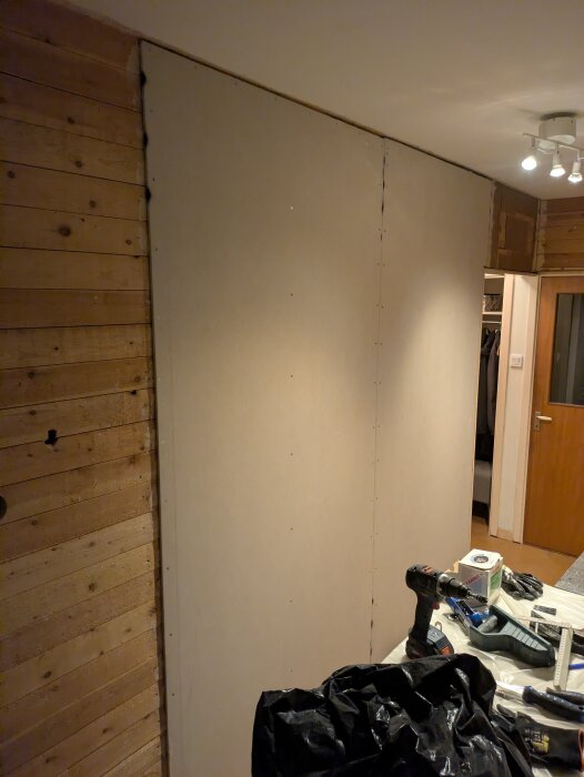 En nyligen uppsatt gipsskiva mot en träribbad vägg, omgiven av verktyg inklusive en borrmaskin och skyddshandskar på ett arbetsbord.