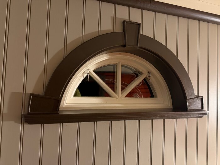 Halvmåneformat fönster med mörkbruna foder färdigmålat, installerat på beige träpanelvägg.