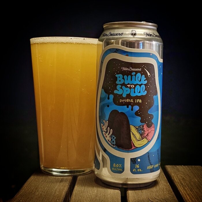 Burk med "Built to Spill" dubbel IPA framför glas med ljust öl på träbord.