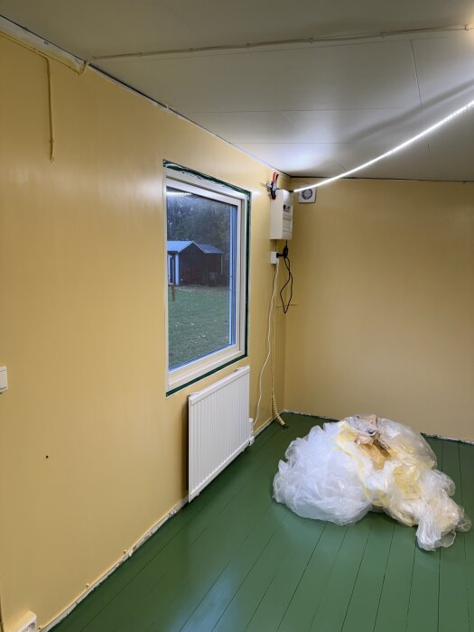 Nymålat grönt trägolv i trädgårdsbod med gula väggar och ett fönster. Plast ligger på golvet, bidragande till renoveringsprojektets avslut.