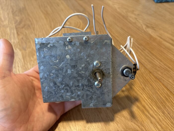 Metalliskt styrkort med kablar och skruvar, hålls i hand mot trägolv.