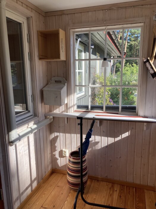 Ljust rum med träpanel, stort fönster, ihopvikt paraply i randig korg, hylla, och brevlåda på vägg. Soligt väder utanför.