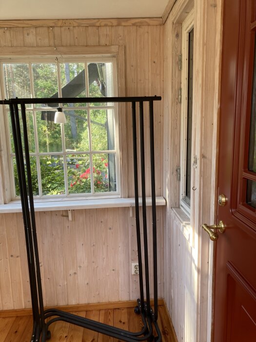 Ett rum med träpanel, stort fönster, och en röd dörr. En svart klädställning står i hörnet. Utanför syns grönska.