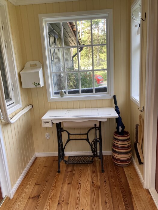 En vitmålad gammal symaskin står vid ett fönster i en hall med ljusa träpaneler. En förvaringslåda med murgrönemönster hänger på väggen.