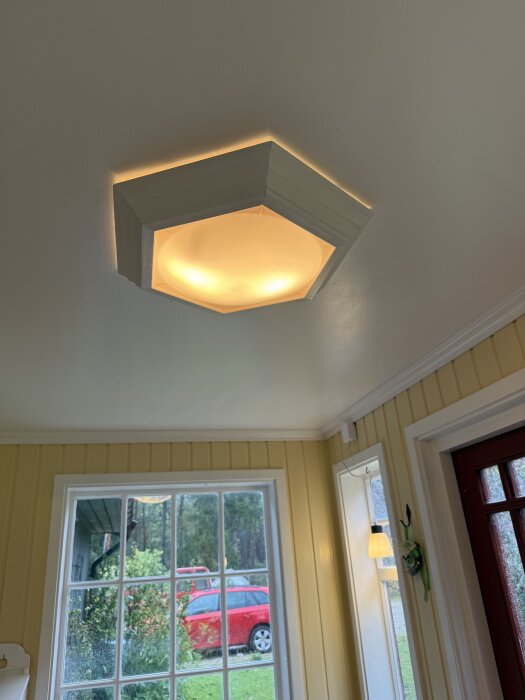 Hexagonal taklampa målad med vit linoljefärg i ett rum med målade träpaneler och naturligt ljus från fönster, röd bil synlig utomhus.